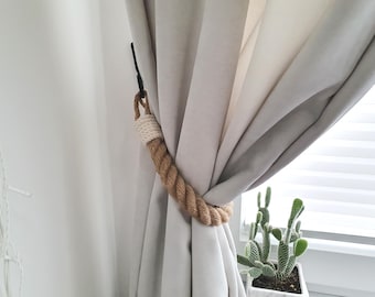 May include: A decorative curtain tieback made of thick, twisted brown rope with a white knitted section. The tieback is holding back a light gray curtain. A black hook attaches the tieback to the wall. A cactus is in a white pot in the background.