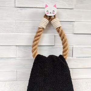 Puede incluir: Un gancho para toallas con forma de gato blanco con orejas rosas y una toalla negra colgando. El gancho está hecho de cuerda y tiene un diseño de cuerda blanca y beige.