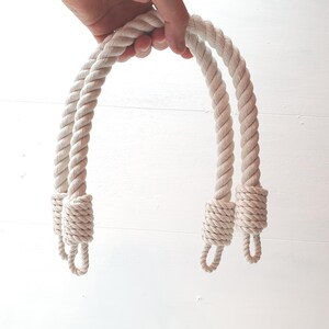 Op de afbeelding: Twee cr&egrave;mekleurige touwhandvatten voor een tas of mand. De handvatten zijn dik en gedraaid, met decoratieve knopen aan de uiteinden. Het touw heeft een neutrale kleur, geschikt voor diverse knutselprojecten.