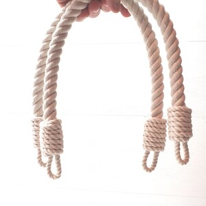 Op de afbeelding: Twee cr&egrave;mekleurige touwhandvatten met luseinden. Het dikke, gedraaide touw wordt tegen een witte achtergrond gehouden. De handvatten zijn aan de basis geknoopt en klaar om aan een tas of mand te worden bevestigd.