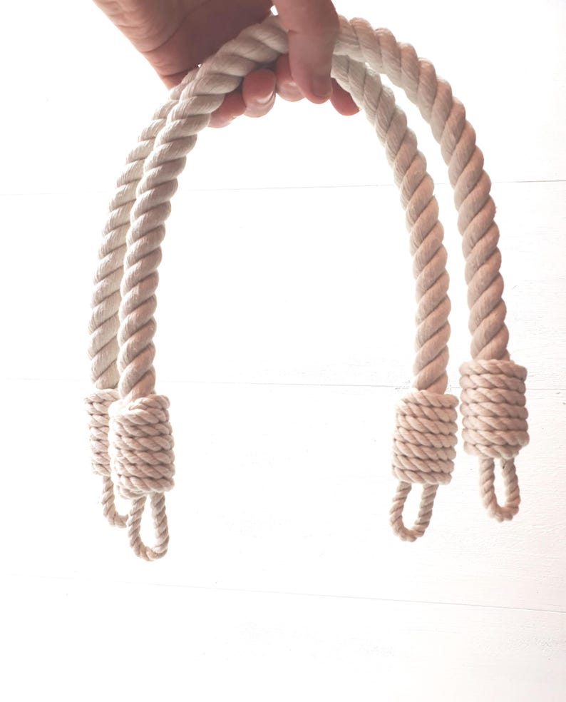 Op de afbeelding: Twee cr&egrave;mekleurige touwhandvatten met luseinden. Het dikke, gedraaide touw wordt tegen een witte achtergrond gehouden. De handvatten zijn aan de basis geknoopt en klaar om aan een tas of mand te worden bevestigd.