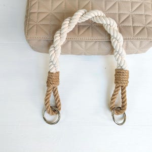 Puede incluir: Bolso beige acolchado con un asa de cuerda blanca trenzada. El asa está asegurada con cuerda marrón y anillos de metal plateado. El bolso está sobre una superficie blanca.