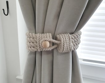 May include: Cream-colored rope curtain tiebacks with a wooden bead closure. The tiebacks are wrapped around light gray curtains. A black hook is visible on the wall in the background. The window blinds are visible in the background.