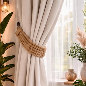May include: Cream-coloured curtains held by a thick, woven rope tieback. The curtains are in front of a window with sheer white curtains. A decorative light fixture and potted plants are visible in the background.