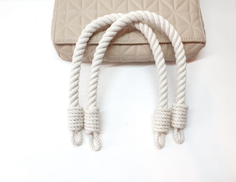 Op de afbeelding: Cr&egrave;mekleurige gewatteerde handtas met witte touwhandvatten. De tas heeft een ruitpatroon en een rechthoekige vorm. De touwhandvatten zijn dik en aan de uiteinden geknoopt. De tas ligt op een witte ondergrond.