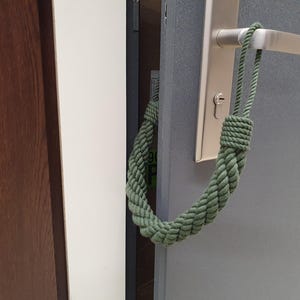 May include: A green braided rope door stop is attached to a silver door handle. The door is slightly ajar, revealing a glimpse of the interior. The door stop is made of a thick, textured rope.