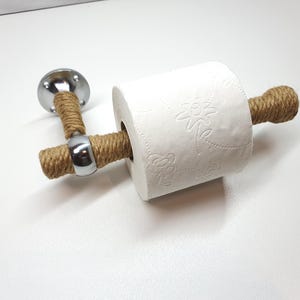May include: A chrome toilet paper holder with a rope wrapped around it. The holder is mounted to a wall and has a roll of toilet paper on it.