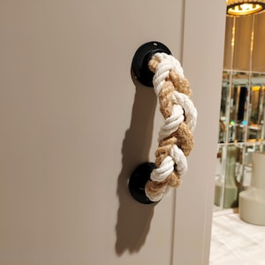 Op de afbeelding: Een witte en bruine touwdeurgreep met zwarte metalen accenten. Het touw is gevlochten en heeft bovenaan een decoratieve knoop.