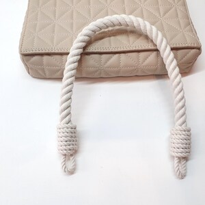 Op de afbeelding: Een beige gewatteerde handtas met een witte touwhandgreep. De tas heeft een geometrisch patroon en een neutrale kleur. De touwhandgreep is dik en aan de uiteinden geknoopt.
