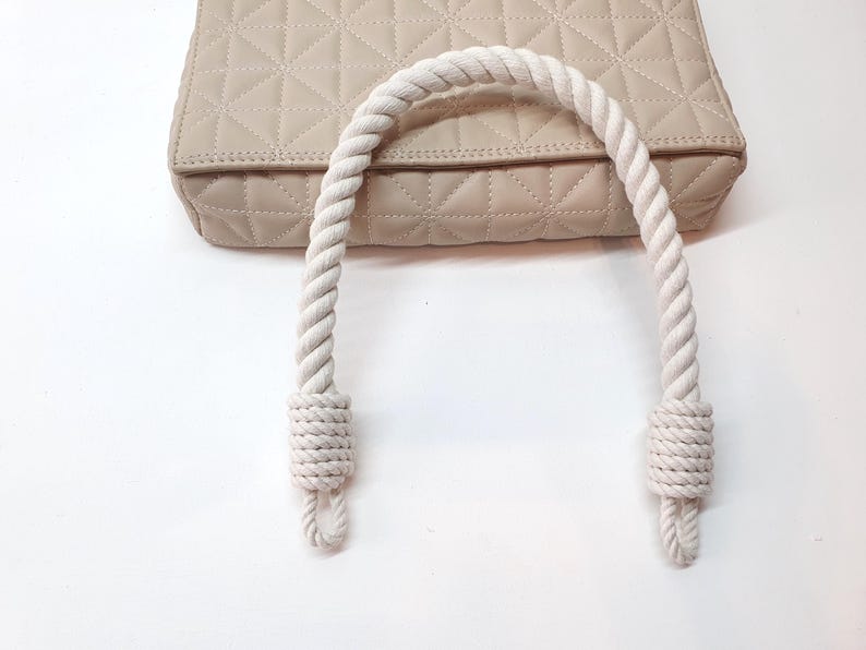 Op de afbeelding: Een beige gewatteerde handtas met een witte touwhandgreep. De tas heeft een geometrisch patroon en een neutrale kleur. De touwhandgreep is dik en aan de uiteinden geknoopt.