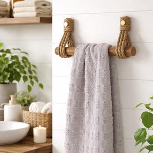 Op de afbeelding: Een houten handdoekenrek met touwaccenten en een grijze handdoek. Het rek is bevestigd aan een witte lambrisering muur. De handdoek heeft een textuurpatroon. Decoratief badkameraccessoire.
