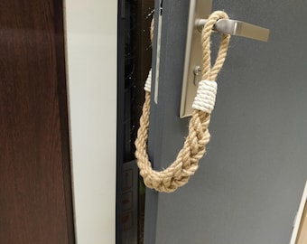 May include: A decorative door handle rope made of braided natural jute fiber. The rope is secured with white twine at the top and bottom. The handle is attached to a gray door with a silver handle.