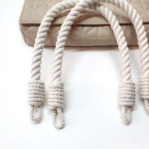 Op de afbeelding: Een beige gewatteerde handtas met dikke, cr&egrave;mekleurige touwhandvatten. De handvatten zijn aan de basis geknoopt en aan de tas bevestigd. De tas heeft een ruitpatroon en een neutrale kleur.