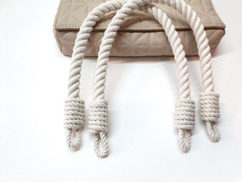 Op de afbeelding: Een beige gewatteerde handtas met dikke, cr&egrave;mekleurige touwhandvatten. De handvatten zijn aan de basis geknoopt en aan de tas bevestigd. De tas heeft een ruitpatroon en een neutrale kleur.