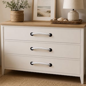 May include: A white wooden dresser featuring three horizontal drawers with thick white rope handles attached by black metal hardware. The dresser has a natural light wood-toned top surface and stands on four short, straight legs.