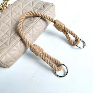 Può includere: Una borsa trapuntata beige con manico in corda. Il manico è realizzato in corda di fibra naturale ritorta, annodata ad ogni estremità e fissata ad anelli in metallo argentato. È visibile anche una tracolla a catena dorata.