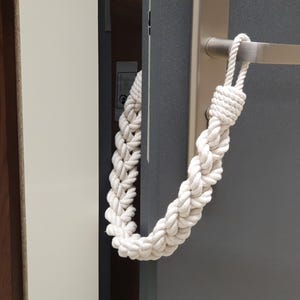 May include: White cotton rope doorstop hanging on a door handle. The rope is braided and has a loop at the top.