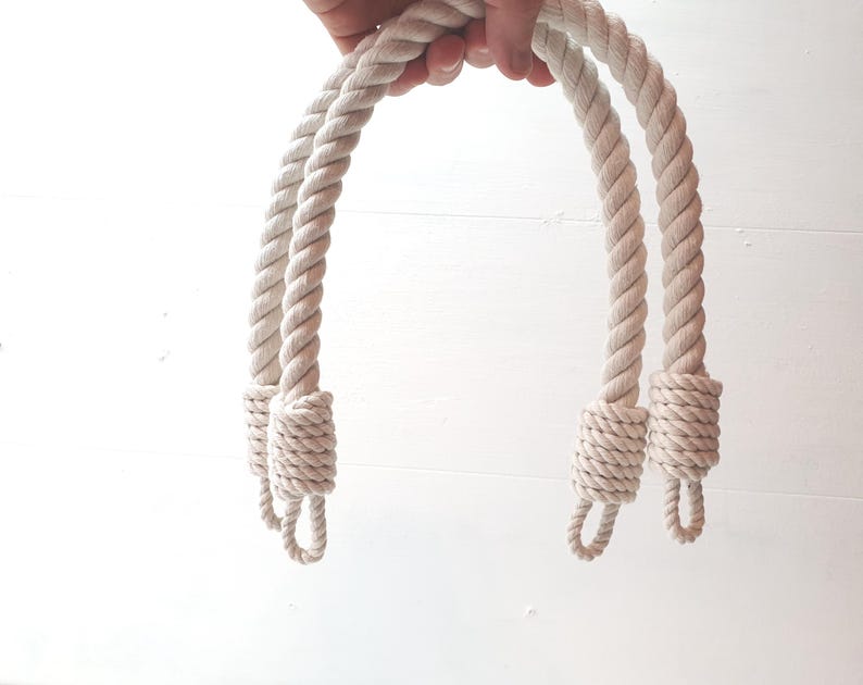 Op de afbeelding: Twee cr&egrave;mekleurige touwhandvatten, tegen een witte achtergrond. De dikke, gedraaide touwen hebben lussen aan de onderkant en gewikkelde secties aan de basis. De natuurlijke kleur van het touw suggereert een nautisch of rustiek thema.