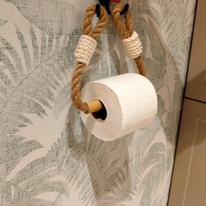May include: A white toilet paper roll is hanging on a rope dispenser with wooden dowel. The dispenser is mounted on a wall with a white and green tropical leaf pattern wallpaper.