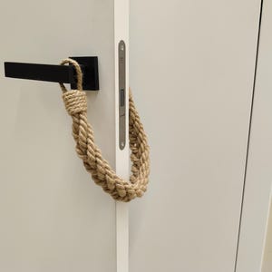 Może przedstawiać: Czarny klamka do drzwi z przymocowanym sznurkowym uchwytem do drzwi. Sznur wykonany jest z naturalnego włókna i jest spleciony w pętlę, tworząc uchwyt.