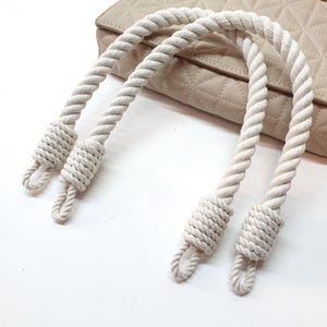Op de afbeelding: Cr&egrave;mekleurige touwhandvatten voor een handtas, met een geknoopt ontwerp aan elk uiteinde. De handvatten zijn gebogen en een beige gewatteerde handtas is zichtbaar op de achtergrond, rustend op een wit oppervlak. Het touw heeft een gedraaide textuur.