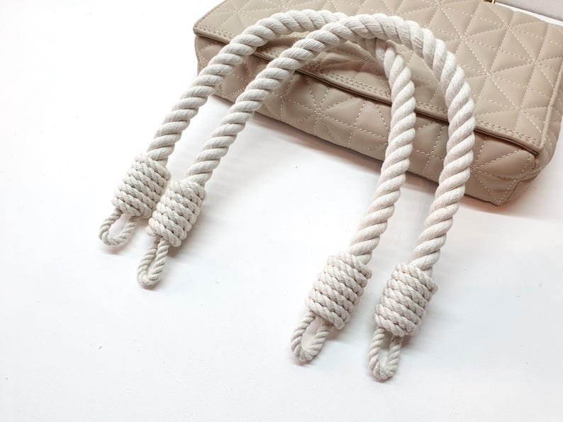 Op de afbeelding: Cr&egrave;mekleurige touwhandvatten voor een handtas, met een geknoopt ontwerp aan elk uiteinde. De handvatten zijn gebogen en een beige gewatteerde handtas is zichtbaar op de achtergrond, rustend op een wit oppervlak. Het touw heeft een gedraaide textuur.
