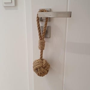 Könnte beinhalten: Ein dekorativer Türstopper aus naturfarbenem Seil. Der Türstopper ist unten geknotet und hat oben eine Schlaufe zum Aufhängen. Er hängt an einem silbernen Türgriff an einer weißen Tür.