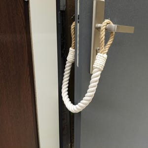 May include: A door stop made of a thick, twisted white rope with tan rope accents, looped around a silver door handle. The door is gray, and the door frame is a dark brown.