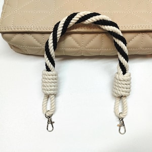 Puede incluir: Un par de asas de cuerda negra y blanca con cierres plateados. Las asas están unidas a un bolso acolchado beige.