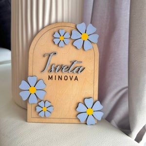 Può includere: Un cartello in legno con il nome "Tsveta MINOVA" in corsivo grigio. Decorato con fiori azzurri con centri gialli. Il cartello ha una parte superiore ad arco ed è in legno chiaro.