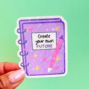 May include: A purple notebook sticker with the text "Create your own FUTURE". The sticker has a spiral binding, a pink pen, and is decorated with hearts, stars, and sparkles.
