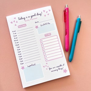 May include: A white daily planner with pink hearts and a pink star. The planner has sections for schedule, date, grateful for, to do, and important. The text "Today is a good day!" is at the top of the planner. The text "You are capable of amazing things!" is at the bottom of the planner.