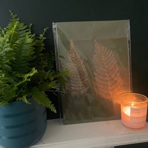May include: A framed print of two fern leaves on a green background. The print is sitting on a white mantelpiece with a candle in front of it.