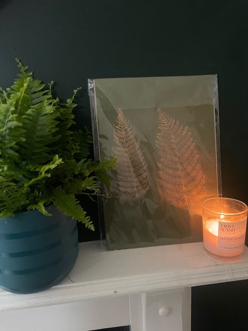May include: A framed print of two fern leaves on a green background. The print is sitting on a white mantelpiece with a candle in front of it.