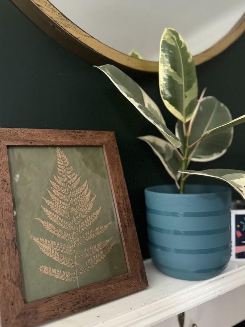May include: A framed print of a gold fern on a green background, with a potted plant with variegated leaves in the background. The plant is in a teal blue planter with white stripes.