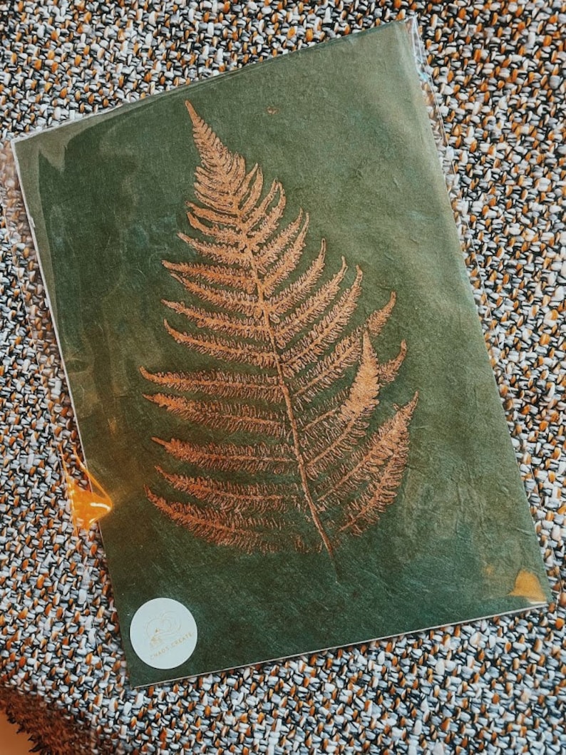 May include: A gold fern leaf pressed onto a dark green background. The leaf is a natural element and is pressed onto a handmade paper. The paper has a textured surface.