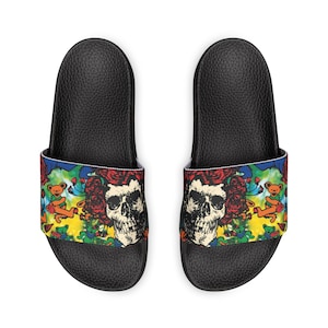 May include: Pair of black slide sandals featuring a colorful design. The design includes a skull, roses, and dancing bears. The sandals have a textured footbed and a wide strap with the graphic print.