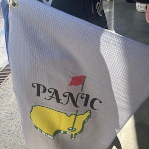 May include: White golf towel with a yellow outline of the United States and the word "PANIC" in black lettering. A red flag is in the upper right corner of the image.