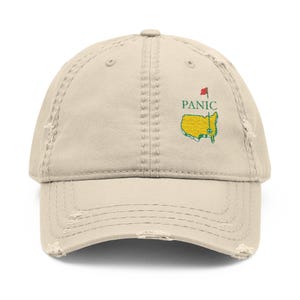 May include: Beige distressed baseball cap with embroidered text and graphic. The text reads "PANIC" above a yellow and green map graphic with a red flag. The cap has a pre-worn look with distressed edges.