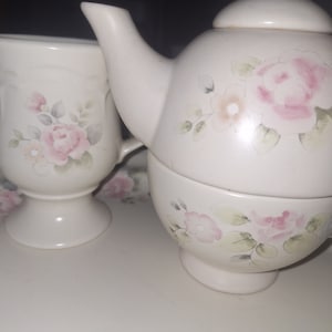 May include: A white ceramic tea set with a floral design. The set includes a teapot, a creamer, and a sugar bowl. The teapot has a lid and a spout. The creamer and sugar bowl have handles. The floral design is in shades of pink and green.