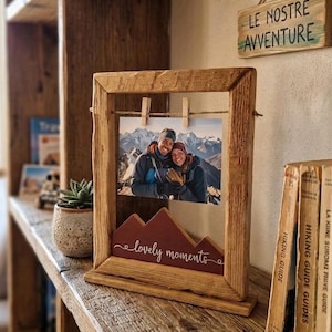 May include: A rustic wooden photo frame with a picture of a couple, held by small clothespins. The frame has a burgundy mountain-shaped accent with the words "lovely moments." A small sign above reads "LE NOSTRE AVVENTURE."
