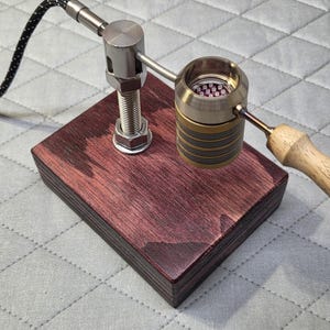 May include: A desktop heating device featuring a dark reddish-brown wooden base, a vertical threaded metal post, and a horizontal arm holding a cylindrical metal heating chamber with a light-coloured wooden handle and a black braided power cord.
