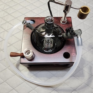 May include: A dark glass water pipe with a wooden base. It features a clear tube, a metal heating element, and a wooden handle. The design includes a central glass chamber and various metal and glass components.