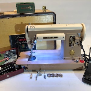 May include: A vintage Singer sewing machine with a white and beige body, a light on the needle, and a foot pedal. The machine is in a beige case with a brown leather handle. The case has the word "Singer" printed on it. There is a manual for the machine that says "Instructions for Singer Sewing Machine".