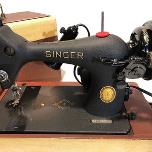 May include: Black Singer sewing machine with a gold logo and a red button on the top. The machine is in a brown wooden case with a beige lining. The case has a gold latch and a gold nameplate that reads "Singer".