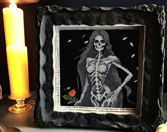 May include: A black framed picture with a cross-stitch design of a skeleton woman with long hair and a single red rose. The picture is on a table with two lit candles and a white pumpkin.