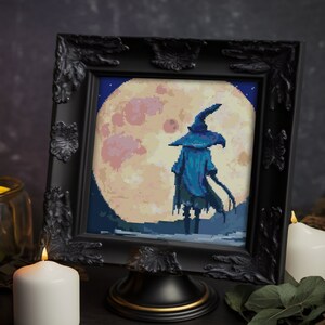 May include: A black framed picture featuring a cross-stitch design of a witch standing before a full moon. The witch is wearing a blue robe and a pointed hat. The moon is yellow with pink and white clouds. The picture is displayed on a black stand.