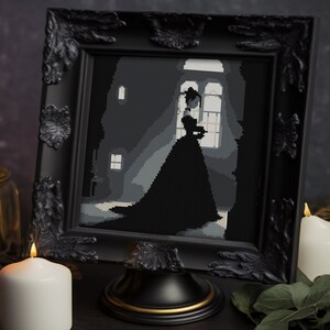 May include: A framed cross-stitch artwork depicting a silhouette of a person in a long gown standing near a window. The black frame has ornate corner details and sits on a black and gold stand. The artwork is in shades of gray and black.
