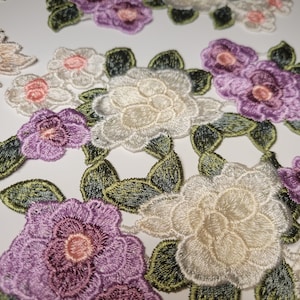 May include: A collection of embroidered fabric flower appliques in shades of white, cream, purple, and green. The appliques feature various flower shapes and sizes, with intricate details and delicate stitching.