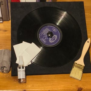 Puede incluir: Un disco de vinilo negro con una etiqueta morada y blanca está centrado en un cuadrado negro. Un pequeño pulverizador, un cepillo, un limpiador de discos gris y cuadrados blancos están dispuestos alrededor del disco.